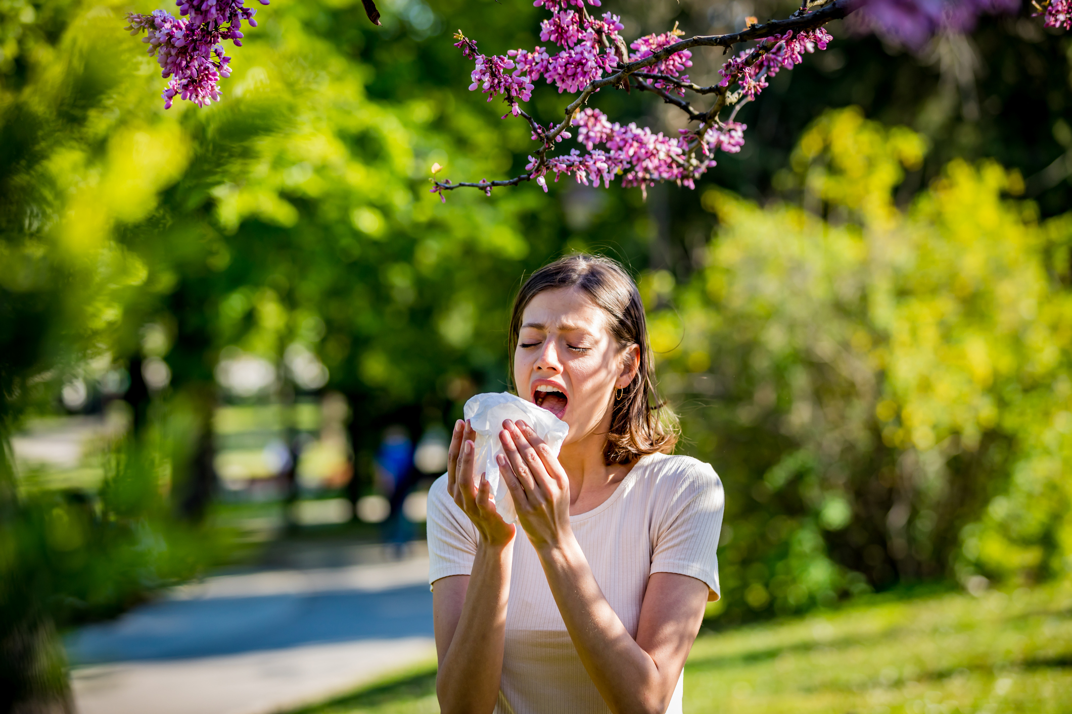 Young pretty woman blowing nose in front of blooming tree. Spring allergy concept