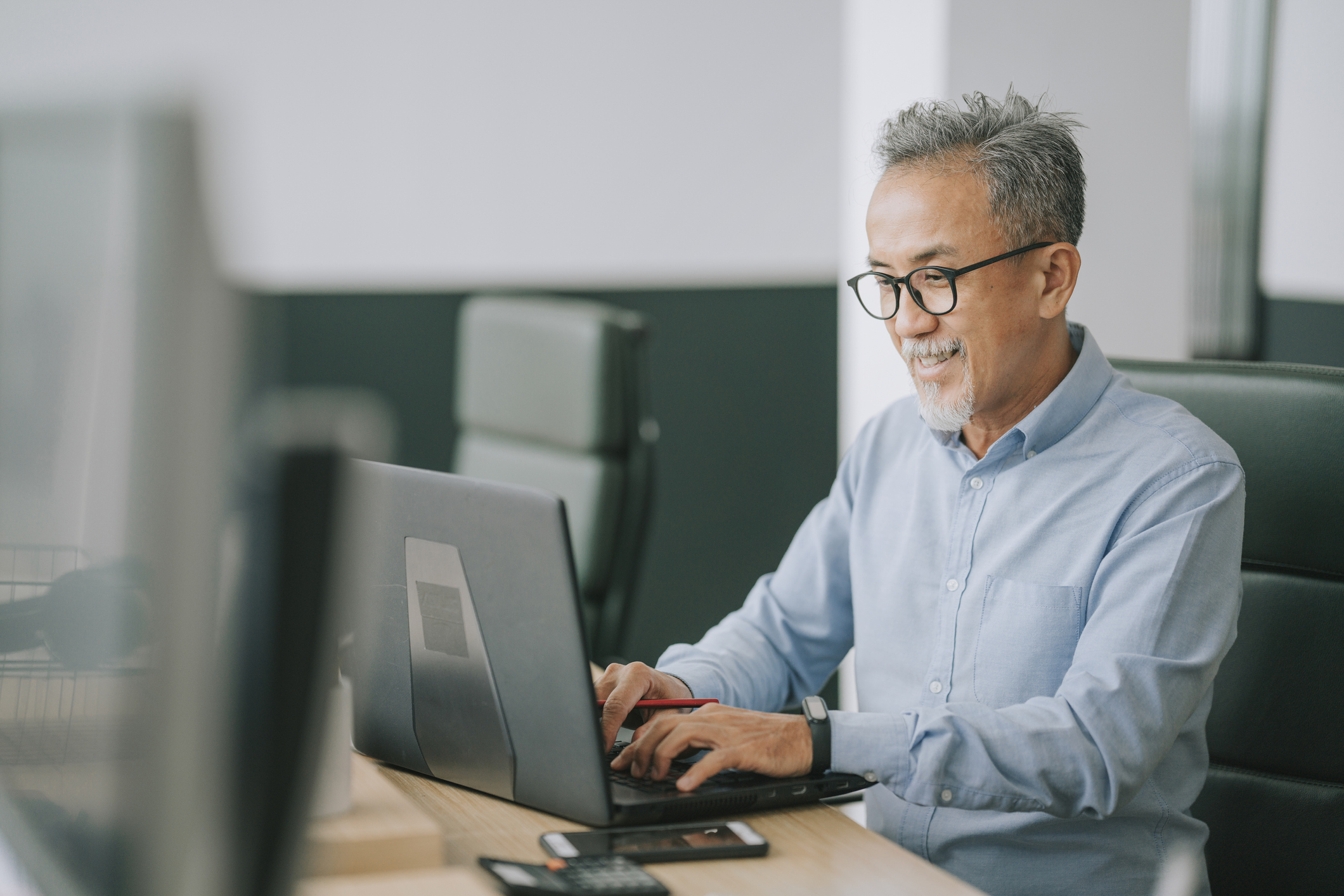 Asian chinese senior man with facial hair using laptop typing working in office open plan